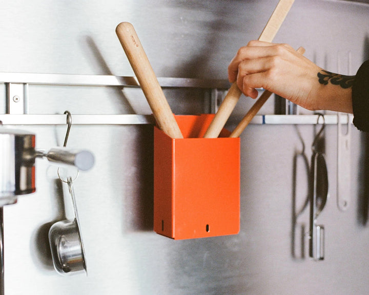 Person placing wooden utensils into an orange container on a kitchen wall.