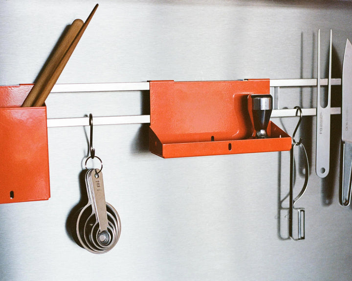 Wall-mounted kitchen organizer with orange shelves and utensils on a white wall.